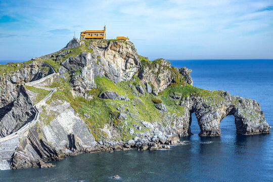 View of the chapel sits atop a rugged islet, connected by a winding stone staircase, amidst the vast blue sea, Gaztelugatxeko Doniene, Euskadi, Spain.