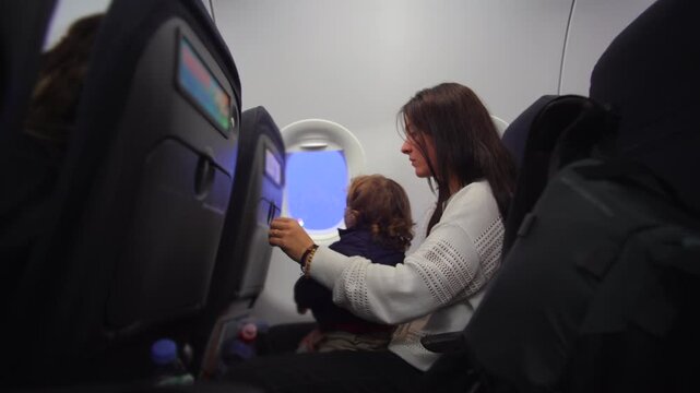 Mother seated on airplane closing tray table and adjusting restless toddler on lap as child reaches toward window evoking patience caregiving effort and preflight tension