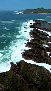 Drone aerial footage following the long jagged rock formations along Karang Taraje Beach, Sawarna, Indonesia. Waves roll in from the open sea creating natural patterns in a clean Vertical perspective.