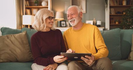 Obraz premium A couple sits together on a couch in a living room looking at a tablet. They talk about life insurance plans and share smiles while discussing their finances.