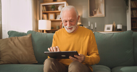 An older man sits on a couch in his living room, smiling as he interacts with a tablet. The room is bright and cozy, with bookshelves and soft furnishings nearby. © Stockphotodirectors
