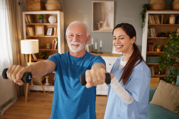 Home health nurse guides senior man in performing exercises with weights in his living room. They...