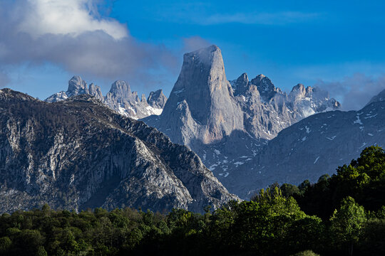 View of the magnificent, cloud-kissed, towering peak standing proudly amidst the rugged, snow-dusted mountains under a vibrant blue sky, Naranjo de Bulnes, Asturias, Spain.