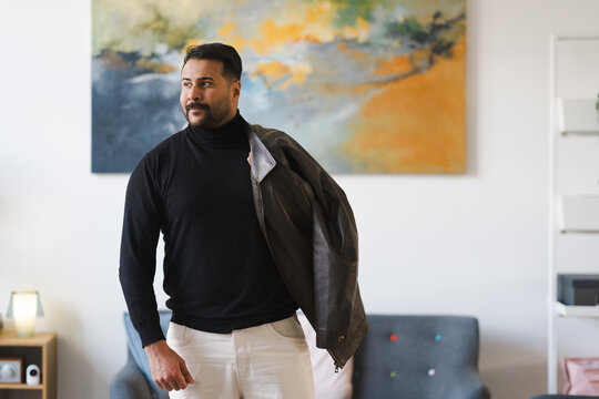 A man in a black turtleneck and light pants stands indoors, holding a brown leather jacket over his shoulder.
