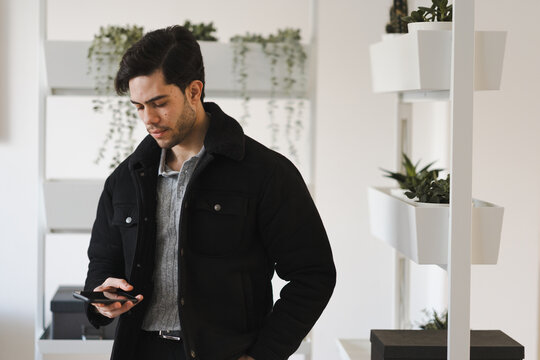 A young man in a black jacket and grey sweater looks down at his phone while standing near a shelf with plants.