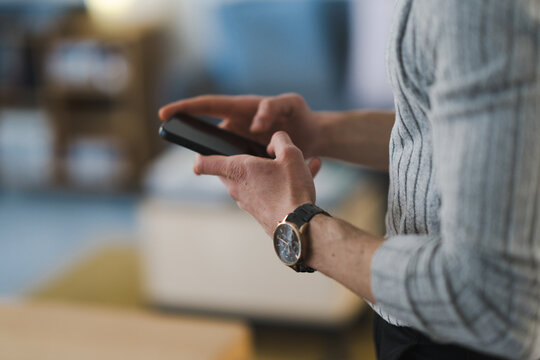 A person wearing a watch uses a smartphone, focusing on the screen. The background is blurred, suggesting an indoor environment.