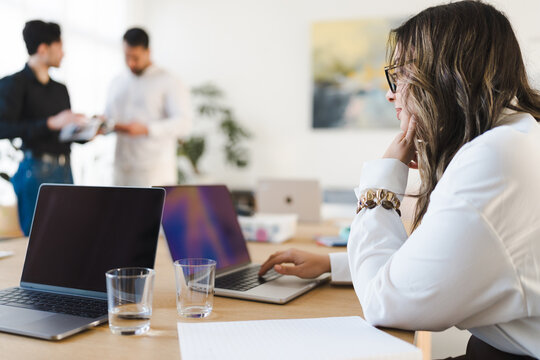 A woman in glasses works on a laptop in an office setting. Two men converse in the background.