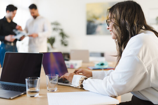 A woman works on her laptop in an office setting while two men discuss documents in the background.