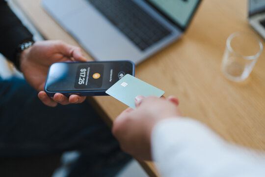 A person holds a smartphone displaying a payment amount while another person taps a credit card to the device, indicating a contactless transaction.