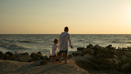 Fototapeta premium Children stroll along the rocky coastline, holding hands. They gaze out at the endless ocean under the warm evening sky, enjoying the feeling of togetherness during the holidays.
