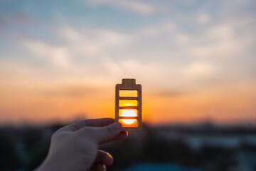 Solar energy concept featuring a hand holding a wooden battery icon against a sunset sky background, representing renewable power, sustainable electricity, and natural energy resource.