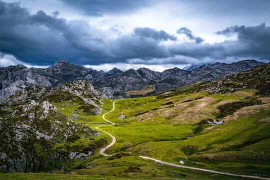 View of a winding path snakes through vibrant green meadows, contrasting with the rugged, grey peaks under a stormy sky, Picos de Europa, Oviedo, Spain.