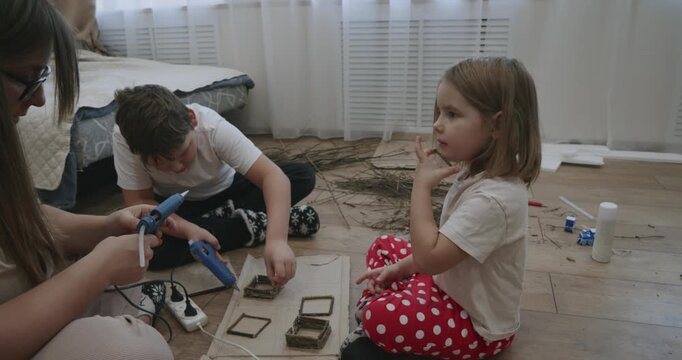 A creative family sits on the floor, crafting together. Mom uses a glue gun to assemble small frames from twigs, and the children help her. The family makes crafts from natural materials.