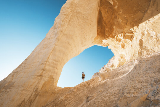 View of a lone figure silhouetted against a vast natural archway carved from pale rock under a clear blue sky, creating a striking contrast, Mangystau, Kazakhstan.