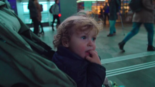 Toddler sitting in stroller at transit hub watching crowd with thoughtful expression suggesting curiosity sensory processing patience and early social awareness