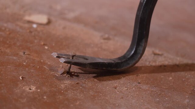 Close-up view of a metal crowbar pulling a rusty nail out of a brown painted wooden surface, showing the force and leverage required for demolition or renovation work in a workshop