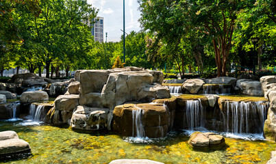 Fototapeta premium Centennial Olympic Park in Atlanta, Georgia. Scenic man-made waterfall flows over rock formations surrounded by green trees and city buildings under a blue sky