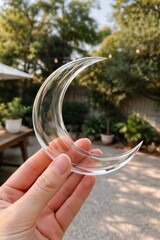 Hand holding clear crescent moon shaped glass outdoors with blurred greenery and patio lights