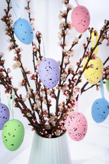 Spring Easter decoration with blooming branches in vase and pastel hanging eggs ornament. Festive composition for holiday greeting, home decor. Selective focus.
