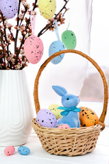 Easter decoration with blue bunny in wicker basket, colorful speckled eggs and spring blossom branches. Selective focus.