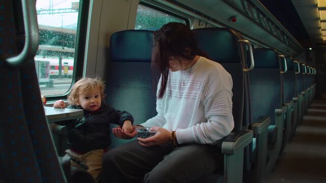 Mother sitting with toddler by train window sharing blueberries during rainy commute showing nurturing care bonding and modern family travel routine