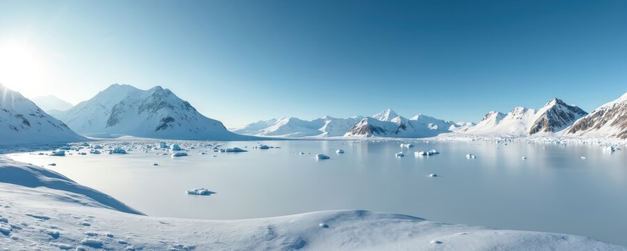 Vast arctic landscape with snow covered mountains and icy water. Small icebergs float in calm frozen bay under clear blue sky. Sun shines brightly.