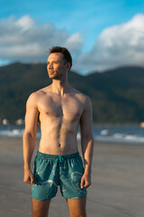 handsome white man with athletic muscular body standing at beach at sunny sunset, attractive male athlete walking at sea or ocean shore