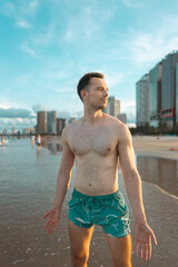 handsome white man with athletic muscular body standing at beach at sunny sunset, attractive male athlete walking at sea or ocean shore