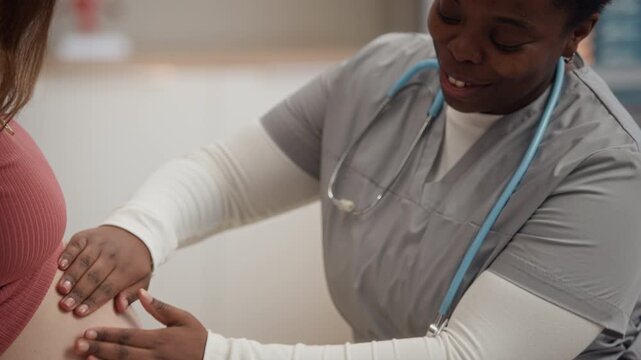 Black female doctor performing prenatal belly examination for Caucasian pregnant woman during routine medical checkup in modern healthcare clinic