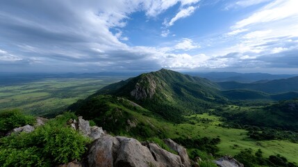 Panoramic vista from a rocky mountain summit overlooking lush green hills and a vast valley under a dramatic cloudy sky