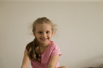 Fototapeta premium Smiling girl in pink shirt sitting by wall, playful afterschool mood with tousled hair, warm expression, natural light, candid childhood moment ideal for parenting and lifestyle