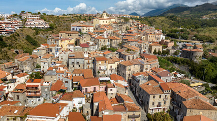 Aerial view of a Mediterranean hillside village, featuring a dense cluster of houses that climb toward a ridge, set against a backdrop of green hills and distant mountain peaks. It's Scalea, Italy.
