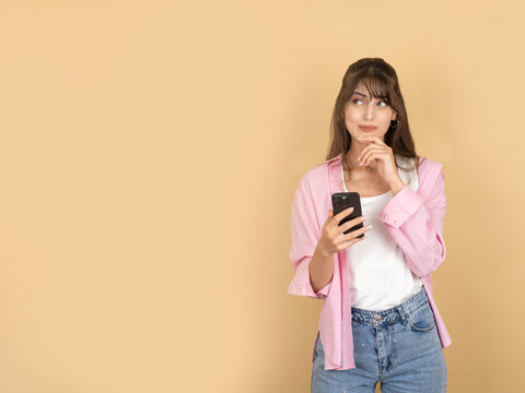 Pensive young woman, portrait of caucasian  20s brunette pensive young woman hold use mobile phone touching chin thinking looking aside copy space isolated on beige background, lifestyle concept.