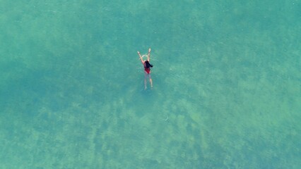 Woman in red swimsuit stands in shallow turquoise water, arms raised in celebration, aerial view showcasing clear ocean and sandy seabed beneath © Irina Flamingo