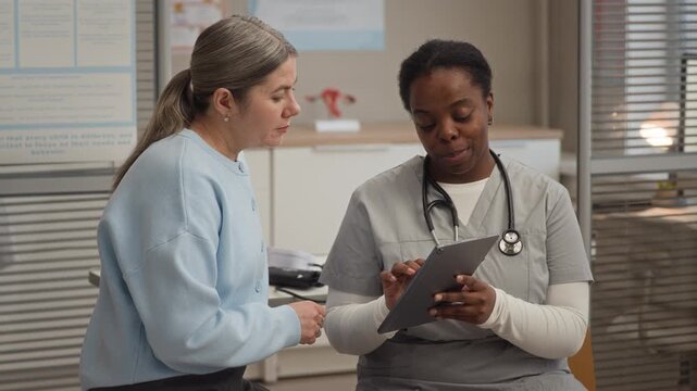Caucasian female patient listening as African American female doctor using digital tablet while discussing examination results and next steps during one on one medical appointment in clinic office