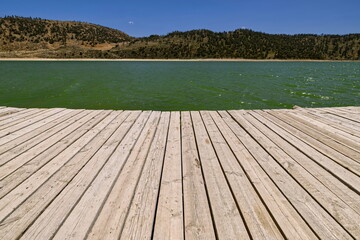 Fototapeta premium Wood boardwalk extending onto the emerald-green water of Lake Aguelmame Sidi Ali, the Middle Atlas mountains in background. Midelt prov.-Morocco-121