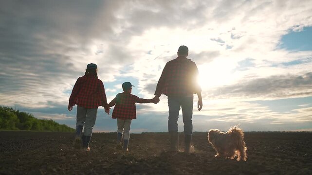 Family of three walking in field with dog. Father holds hands with boy and girl. Rubber boots step into rich soil. Dog walks near boots. Peaceful moment of family walking with child in field.
