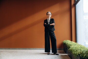 Businesswoman in black suit stands confidently with arms crossed in modern office space featuring a warm brown wall and green plants along the floor © standret