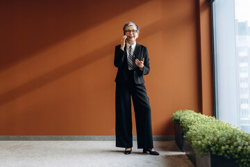 Senior Asian businesswoman in black suit and tie stands confidently in modern office space with orange wall and indoor plants, showcasing professional attire and environment © standret