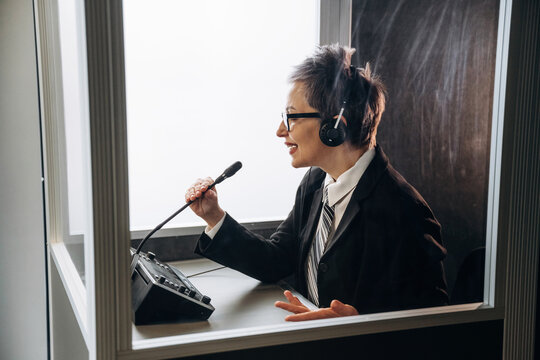 Female interpreter wearing headset speaks into microphone inside soundproof booth, with control panel and glass walls visible in a modern business office setting