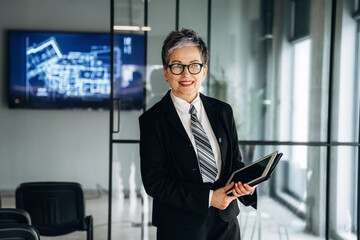 Fototapeta premium Businesswoman in formal attire holding a notebook stands confidently in a modern office with a digital screen displaying architectural plans in the background