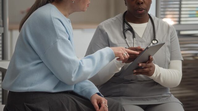 Cropped shot of diverse female doctor and patient reviewing medical test results on digital tablet while discussing diagnosis and next steps during clinic consultation