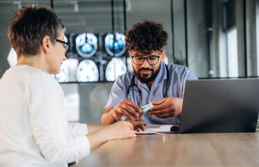 Male healthcare professional in scrubs discusses treatment options with female patient in modern medical office, laptop and medical images visible in the background