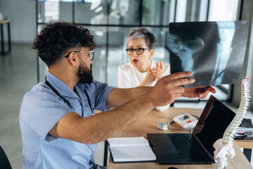Fototapeta premium Male healthcare professional in blue shirt discusses X-ray results with female patient in a modern office setting, laptop and anatomical model visible on the table