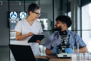 Fototapeta premium Female healthcare professional in white scrubs discusses findings with male colleague in blue scrubs, laboratory setting with microscope and medical imaging in background