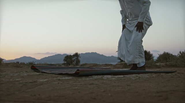Man in white robe praying on a colorful mat in a serene desert landscape at sunset with mountains in the background