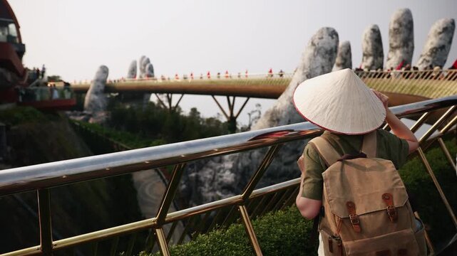 Young woman tourist in conical Vietnamese hat with backpack traveling on golden bridge in Da Nang, Vietnam..