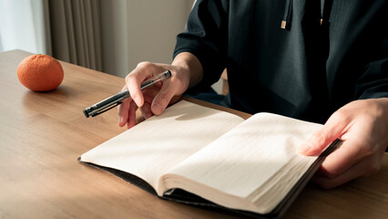ノートに書き物をする男性の手元 思考の整理 ジャーナリング 丁寧な暮らし / Hands writing in notebook with pen and orange on table, journaling and planning concept