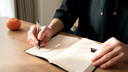 ノートに書き物をする男性の手元 思考の整理 ジャーナリング 丁寧な暮らし / Hands writing in notebook with pen and orange on table, journaling and planning concept