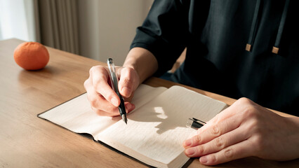 ノートに書き物をする男性の手元 思考の整理 ジャーナリング 丁寧な暮らし / Hands writing in notebook with pen and orange on table, journaling and planning concept
